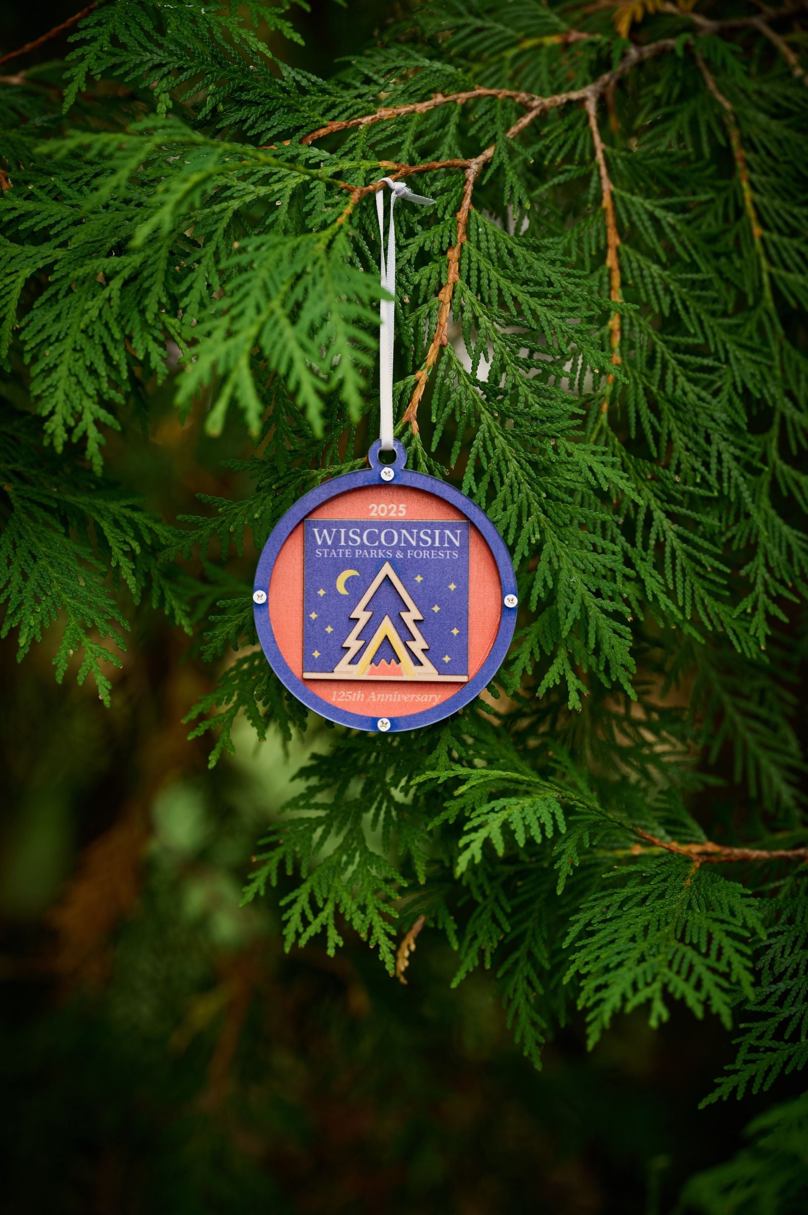 A commemorative blue and orange ornament marking the 125th anniversary of the Wisconsin State Parks System in 2025. The design features a stylized campfire nested inside a tent inside an evergreen tree with a starry sky and crescent moon in the background.