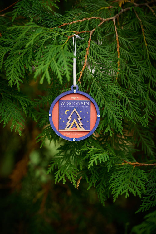 A commemorative blue and orange ornament marking the 125th anniversary of the Wisconsin State Parks System in 2025. The design features a stylized campfire nested inside a tent inside an evergreen tree with a starry sky and crescent moon in the background.